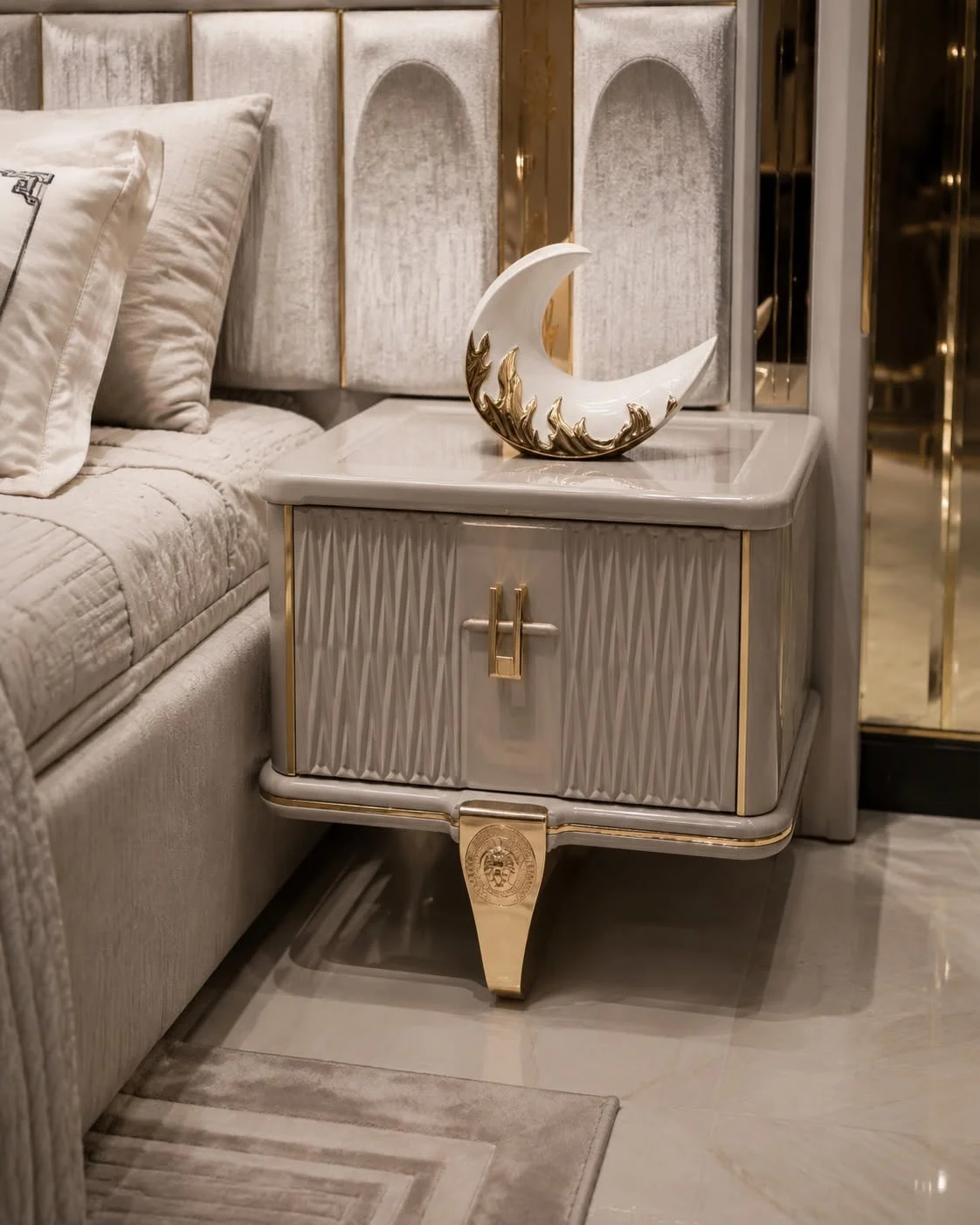 Close-up of the Versace cream nightstand with textured drawer front, gold-tone handle and tapered decorative leg beside the upholstered bed